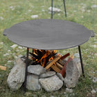 Round metal fire pit on stones with flames and wood inside, surrounded by grass and rocks.
