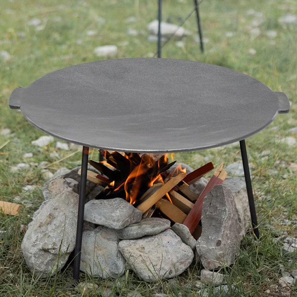 Round metal fire pit on stones with flames and wood inside, surrounded by grass and rocks.