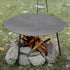 Round metal fire pit on stones with flames and wood inside, surrounded by grass and rocks.